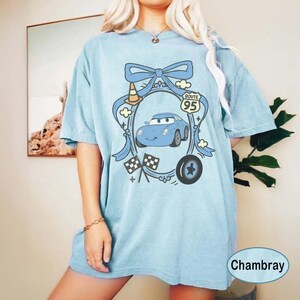 May include: Light blue t-shirt featuring a cartoon car design with a Route 95 sign, checkered flags, and a tire. The design is framed by a blue bow and clouds. The shirt is labeled "Chambray".