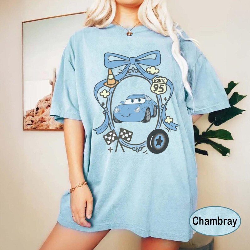 May include: Light blue t-shirt featuring a cartoon car design with a Route 95 sign, checkered flags, and a tire. The design is framed by a blue bow and clouds. The shirt is labeled "Chambray".
