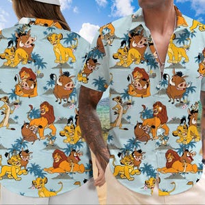 Puede incluir: Una camisa de manga corta con botones azul y blanca con un patrón repetido de leones, hienas y jabalíes de dibujos animados. La camisa tiene cuello y un bolsillo en el lado izquierdo.