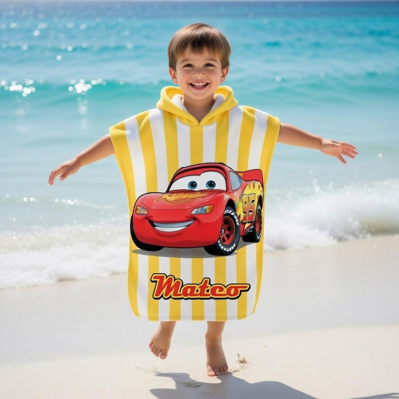 K&ouml;nnte beinhalten: Ein gelb-wei&szlig; gestreiftes Kapuzen-Badetuch mit einem roten Cartoon-Rennwagen mit der Nummer 95. Auf dem Handtuch steht der Name "Mateo". Das Kind steht an einem Sandstrand mit dem Meer im Hintergrund.