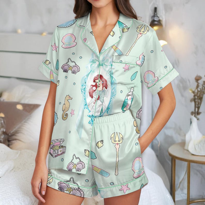 May include: Light green pajama set featuring a print of Ariel from The Little Mermaid, seashells, and other aquatic elements. The short-sleeved top has a portrait of Ariel. The shorts have an elastic waistband. The set is made of a silky material.