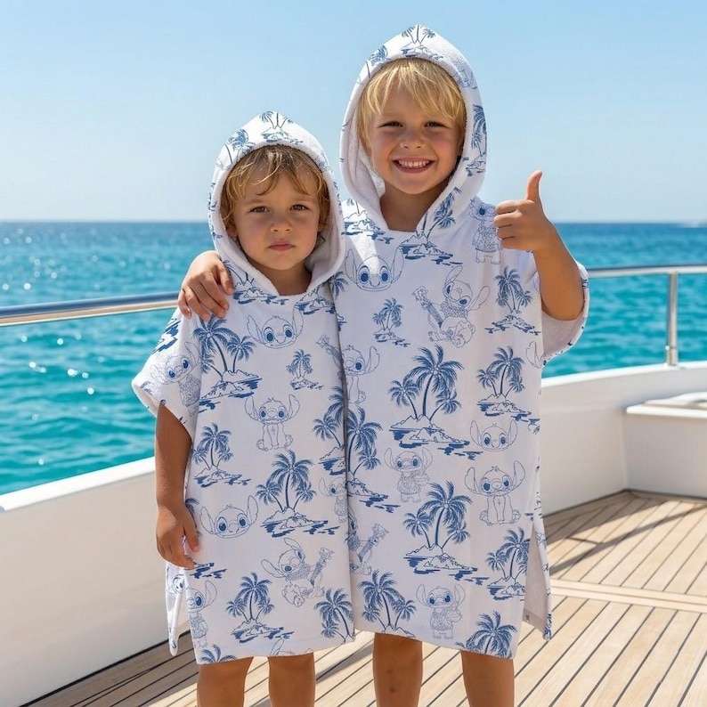 May include: Two white hooded poncho towels featuring a repeating blue print of the character Stitch from Lilo and Stitch, interspersed with palm trees and island scenery. The hooded towels have a loose, sleeveless fit and reach mid-thigh length.