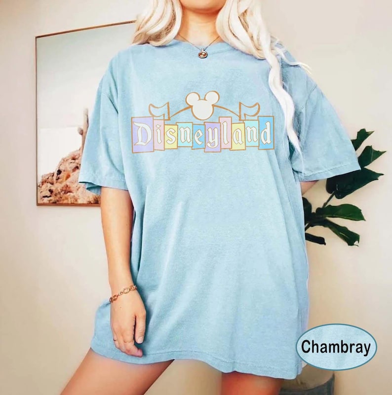 May include: Pale blue t-shirt with the word "Disneyland" in pastel colours, featuring the iconic Mickey Mouse ears. The shirt has a relaxed fit and short sleeves. The word "Chambray" is in a white oval on the bottom right.