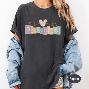 May include: Charcoal grey t-shirt with the word "Disneyland" in pastel shades, featuring a Mickey Mouse head and flags. The shirt has rolled-up sleeves and a relaxed fit, suitable for a day out.