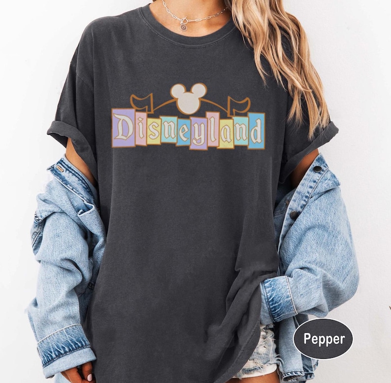 May include: Charcoal grey t-shirt with the word "Disneyland" in pastel shades, featuring a Mickey Mouse head and flags. The shirt has rolled-up sleeves and a relaxed fit, suitable for a day out.