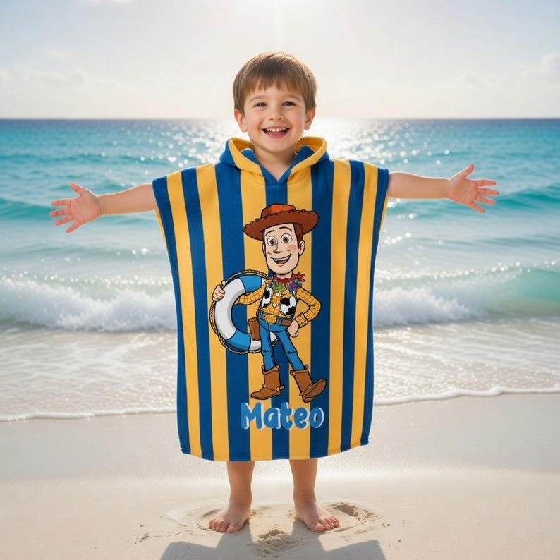May include: A hooded beach towel featuring a cartoon image of Woody from Toy Story, with blue and yellow vertical stripes. The towel has the name "Mateo" printed on it. The child is standing on a sandy beach with the ocean in the background.
