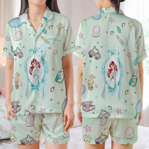 May include: Light green pajama set featuring a print of Ariel from The Little Mermaid. The short-sleeved top and shorts have a repeating pattern of seashells, seahorses, and other aquatic elements. The set includes a large framed image of Ariel.