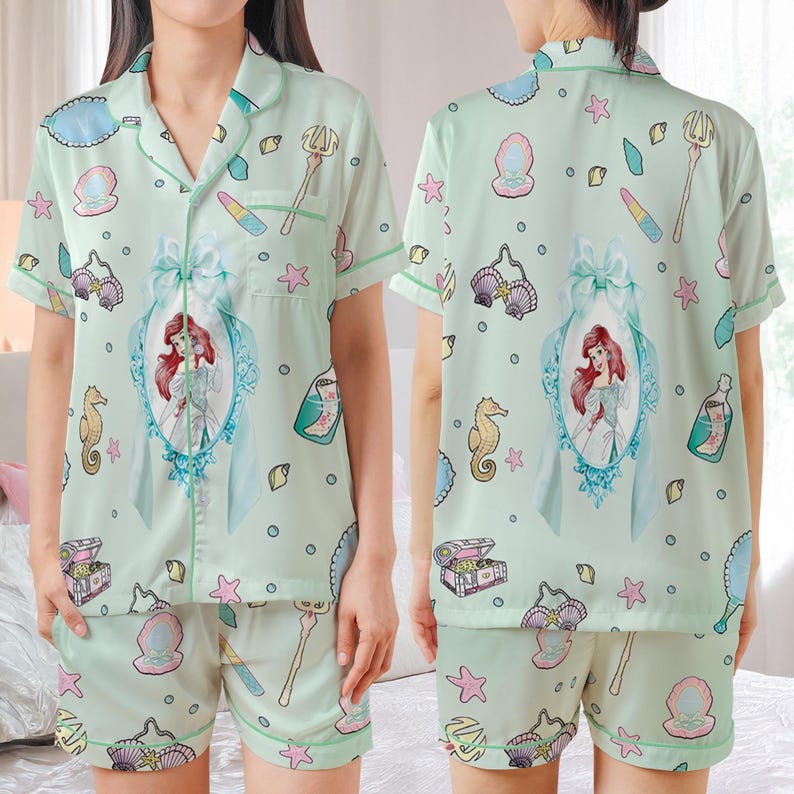 May include: Light green pajama set featuring a print of Ariel from The Little Mermaid. The short-sleeved top and shorts have a repeating pattern of seashells, seahorses, and other aquatic elements. The set includes a large framed image of Ariel.