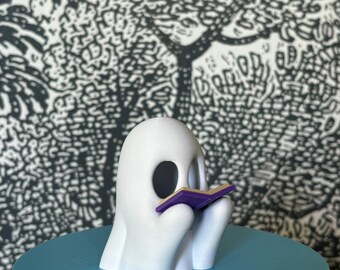 Op de afbeelding: Een wit spookfiguur dat een paars boek leest. Het spook zit op een lichtblauw rond oppervlak.
