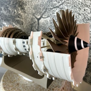 May include: A white and brown model of a jet engine with a cutaway view of the internal components. The engine is mounted on a base and has a fan blade assembly at the front.