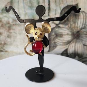 May include: A gold-tone mouse brooch with red rhinestone accents. The brooch is displayed on a black metal stand.