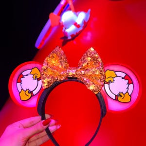 May include: A black headband with two red circular ears featuring a cartoon duck design. A large gold sequined bow sits atop the headband. The background is red, and a glowing duck-shaped light is visible.
