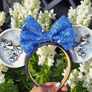 May include: White Minnie Mouse ears with a blue sequin bow. The ears feature a 3D Olaf design with snowflakes.