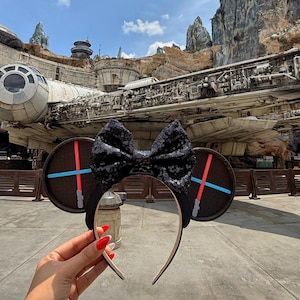 Puede incluir: Diadema con lazo negro con lentejuelas y orejas redondas con diseños de sables de luz rojos y azules. La diadema se sostiene frente a un fondo temático de Star Wars con el Halcón Milenario.