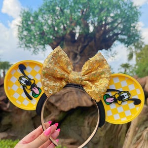 May include: A Minnie Mouse ears headband with yellow and black details and a gold sequined bow. The ears have a yellow and white chequered pattern with a Pluto graphic. The headband is black.