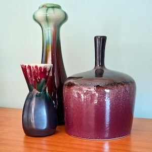 Op de afbeelding: Drie keramische vazen van verschillende vormen en maten. De hoogste vaas is slank met een bolvormige bovenkant, met groene, bruine en oranje tinten. Een kleinere vaas heeft een uitlopende bovenkant met rode en groene glazuur. De grootste vaas is diep bordeaux van kleur.