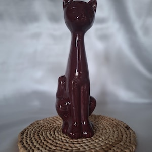 Op de afbeelding: Een glanzend, donkerbordeaux keramieken kattenbeeldje met een langgerekte nek en een zittende houding. De kat heeft puntige oren, een glad oppervlak en een strakke vorm, wat een gestileerde weergave van een kat in een verticale positie vastlegt.