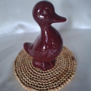 Op de afbeelding: Een glanzend, diepbordeauxrood keramieken eendenbeeldje met een glad, afgerond lichaam, een gedefinieerde snavel en een kleine staart. De eend is staand vormgegeven met zichtbare vleugeldetails aan de zijkanten en een stevige, platte basis.
