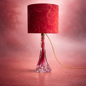 Op de afbeelding: Decoratieve tafellamp met een rode lampenkap en een heldere, gedraaide glazen voet. De lampenkap heeft een textuurpatroon. De lamp staat op een roze oppervlak en heeft een goudkleurig snoer.