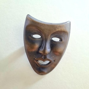 Op de afbeelding: Een bronzen masker met een menselijk gezichtsontwerp. Het masker heeft ovale ooggaten, een prominente neus en een lachende mond. Het oppervlak van het masker heeft een verweerde uitstraling, met donkerdere schaduwen rond de ogen en mond.