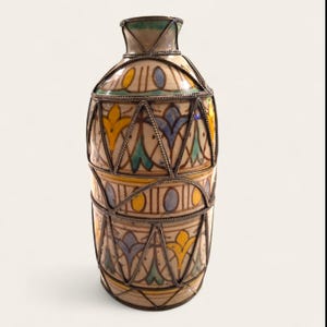 Op de afbeelding: Een hoge, decoratieve keramische vaas met een crèmekleurige basis, versierd met gele, blauwe en groene bloempatronen. De vaas is omhuld door een zilverkleurig metalen frame. Metalen banden scheiden de secties.