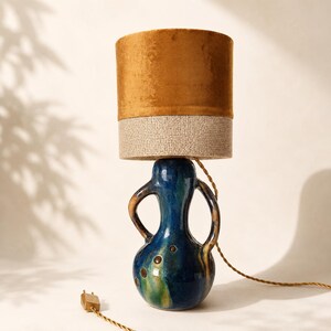 Op de afbeelding: Een tafellamp met een glanzende, diepblauwe keramische voet met twee gebogen handvatten en kleine ronde uitsparingen. De cilindrische lampenkap is verdeeld in twee secties, met een gladde, mosterdkleurige fluwelen bovenkant en een gestructureerde, beige stoffen onderkant.