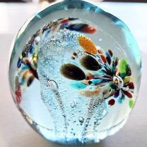 Op de afbeelding: Een heldere glazen presse-papier met kleurrijke, abstracte ontwerpen. De presse-papier heeft een ronde vorm en bevat een mix van blauw, oranje, groen en bruin. Kleine belletjes zijn zichtbaar in het glas, wat bijdraagt aan de decoratieve uitstraling.