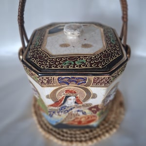 Op de afbeelding: Een zeshoekige keramische houder met een craquelé glazuurafwerking, voorzien van een deksel met een kleine knop en een gebogen gevlochten handvat. De buitenkant is versierd met kleurrijke geschilderde figuren en gouden accenten op een witte en zwarte achtergrond.