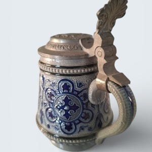 Op de afbeelding: Een vintage keramische bierpul met een scharnierend metalen deksel en handvat. De pul heeft een blauw en wit bloemenpatroon met een decoratieve metalen handgreep en deksel. De handgreep en het deksel zijn zilverkleurig. De pul is een traditionele stijl.