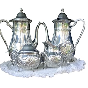 Op de afbeelding: Een zilveren theeservies met sierlijke details, waaronder twee theepotten, een suikerpot en een melkkan. De set wordt gepresenteerd op een witte, getextureerde doek, die het reflecterende oppervlak en het klassieke ontwerp benadrukt.