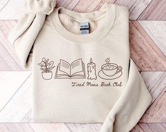 Può includere: Una felpa beige con girocollo e maniche lunghe. La parte anteriore presenta un disegno a tratto marrone di una pianta in vaso, un libro aperto, una candela e una tazza e piattino. Le parole "Tired Moms Book Club" sono stampate sotto.