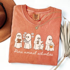May include: A terracotta-colored short-sleeve t-shirt featuring a graphic of five ghosts engaged in various tasks with the text "Para normal activities" printed below. The shirt has a crew neckline and a Comfort Colors brand label at the collar.