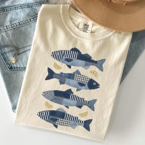 May include: Cream-colored t-shirt featuring four fish designs in shades of blue, with striped and plaid patterns. Each fish is accompanied by a lemon slice. The shirt is folded and placed with a hat and jeans.
