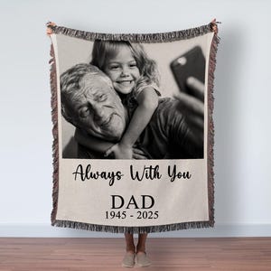 May include: A woven throw blanket with a black and white photo of a man and a child. The blanket has a cream-colored background with the words "Always With You" and "DAD 1945-2025" printed on it. The blanket has a fringed edge.
