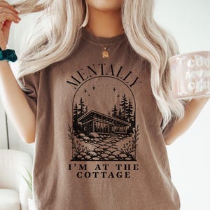 May include: A brown t-shirt with the text "MENTALLY I'M AT THE COTTAGE" in black. The graphic features a cabin, trees, and stars. The person is also wearing a teal scrunchie and holding a pink mug.