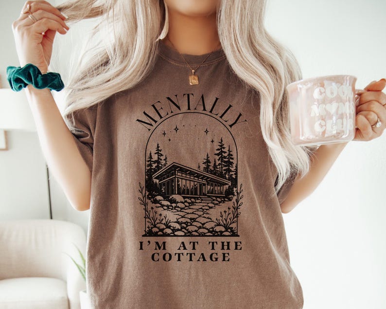 May include: A brown t-shirt with the text "MENTALLY I'M AT THE COTTAGE" in black. The graphic features a cabin, trees, and stars. The person is also wearing a teal scrunchie and holding a pink mug.