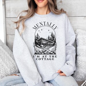 May include: A light gray sweatshirt with the text "MENTALLY I'M AT THE COTTAGE" above a black and white illustration of a cottage surrounded by trees and a stone path. The sweatshirt is a crew neck style.