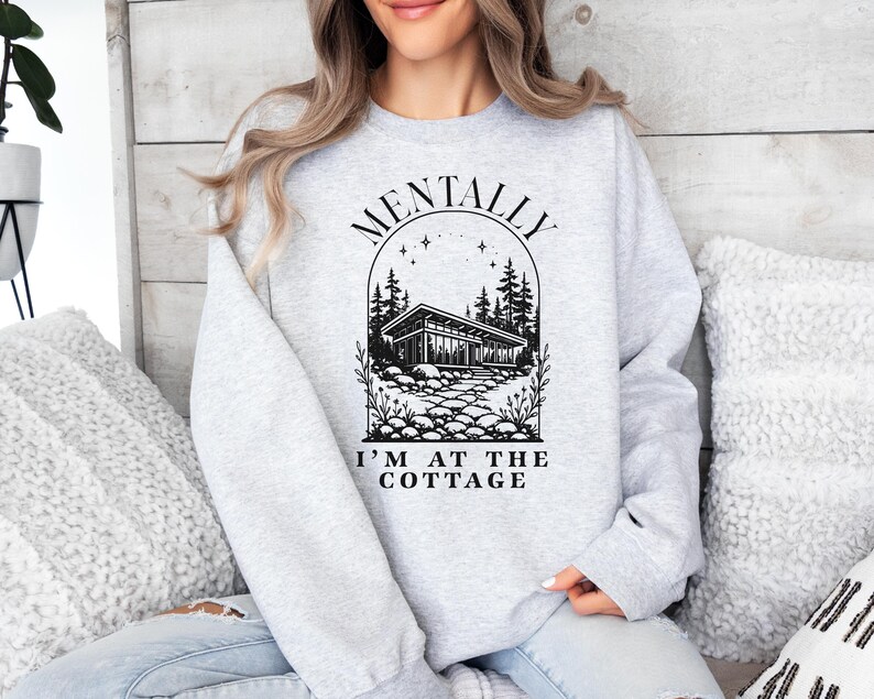 May include: A light gray sweatshirt with the text "MENTALLY I'M AT THE COTTAGE" above a black and white illustration of a cottage surrounded by trees and a stone path. The sweatshirt is a crew neck style.
