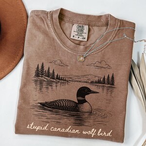 May include: A brown Comfort Colors t-shirt featuring a black and white illustration of a loon on a lake with trees and mountains. The text "stupid canadian wolf bird" is printed at the bottom. A silver necklace with a small pendant is on the shirt.