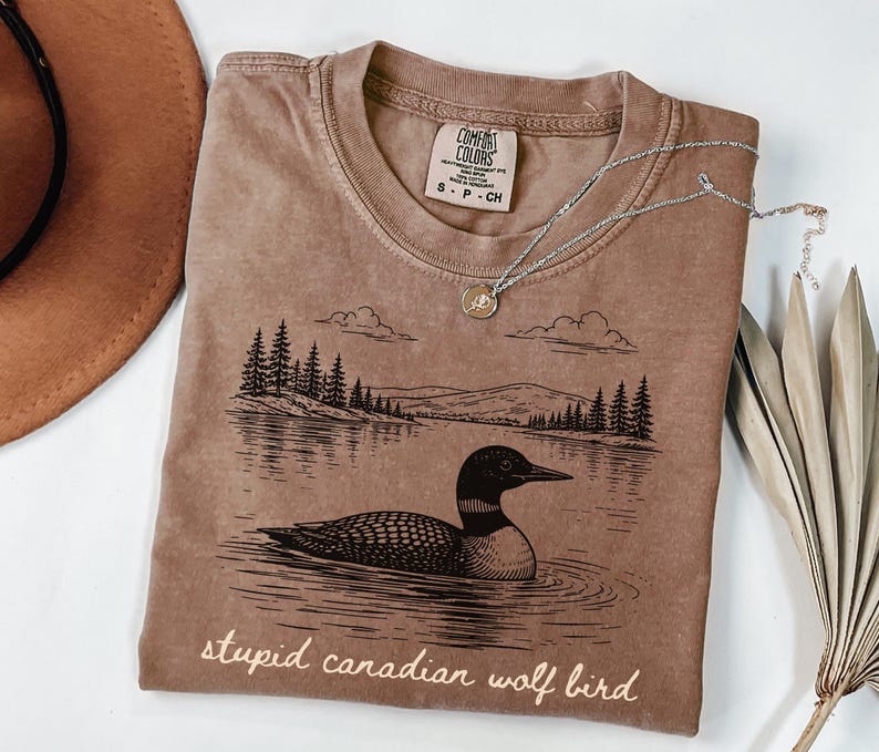 May include: A brown Comfort Colors t-shirt featuring a black and white illustration of a loon on a lake with trees and mountains. The text "stupid canadian wolf bird" is printed at the bottom. A silver necklace with a small pendant is on the shirt.