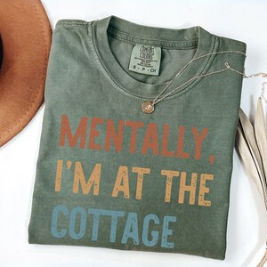 May include: Olive green t-shirt with the text "MENTALLY, I'M AT THE COTTAGE" in orange, tan, and blue. A gold necklace and a brown hat are also visible. The shirt is made of a soft material.