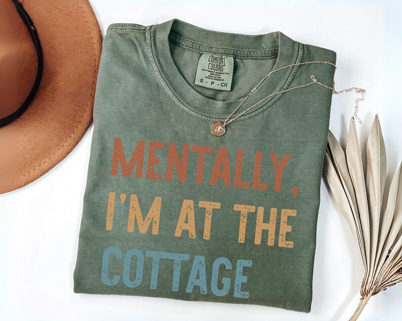 May include: Olive green t-shirt with the text "MENTALLY, I'M AT THE COTTAGE" in orange, tan, and blue. A gold necklace and a brown hat are also visible. The shirt is made of a soft material.