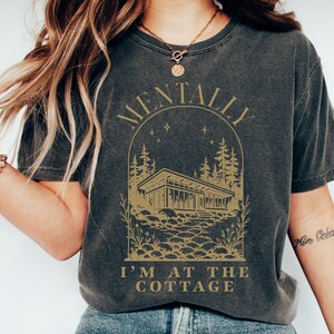 May include: Dark gray t-shirt with a gold graphic featuring a house, trees, and the text "MENTALLY I'M AT THE COTTAGE". The design is enclosed in an arch shape. The shirt has a vintage, worn look.