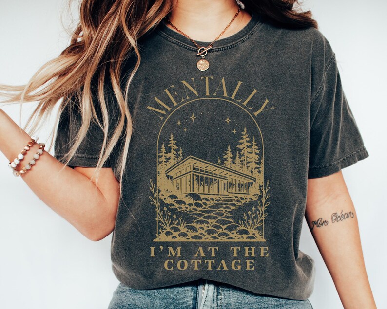 May include: Dark gray t-shirt with a gold graphic featuring a house, trees, and the text "MENTALLY I'M AT THE COTTAGE". The design is enclosed in an arch shape. The shirt has a vintage, worn look.