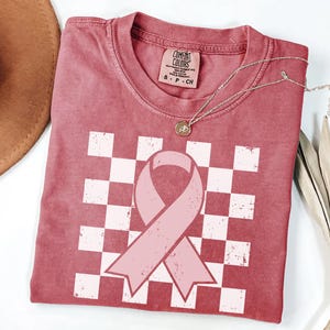 May include: A dusty rose t-shirt featuring a pink ribbon design over a white and pink chequered pattern. The shirt has a crew neck and short sleeves. A silver necklace with a pendant is draped across the shirt.