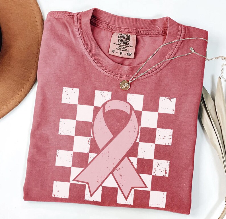 May include: A dusty rose t-shirt featuring a pink ribbon design over a white and pink chequered pattern. The shirt has a crew neck and short sleeves. A silver necklace with a pendant is draped across the shirt.
