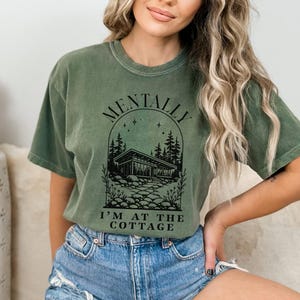May include: Olive green t-shirt with the text "MENTALLY I'M AT THE COTTAGE" in black. The graphic features a house, trees, and stars. The shirt is made of a soft material and has a relaxed fit.