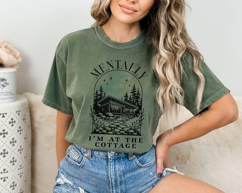 May include: Olive green t-shirt with the text "MENTALLY I'M AT THE COTTAGE" in black. The graphic features a house, trees, and stars. The shirt is made of a soft material and has a relaxed fit.