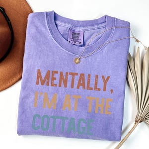 May include: A lavender Comfort Colors t-shirt with the phrase "MENTALLY, I'M AT THE COTTAGE" printed in brown, orange, and green. A gold necklace with a pendant and a brown hat are next to the shirt.