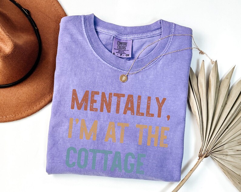 May include: A lavender Comfort Colors t-shirt with the phrase "MENTALLY, I'M AT THE COTTAGE" printed in brown, orange, and green. A gold necklace with a pendant and a brown hat are next to the shirt.